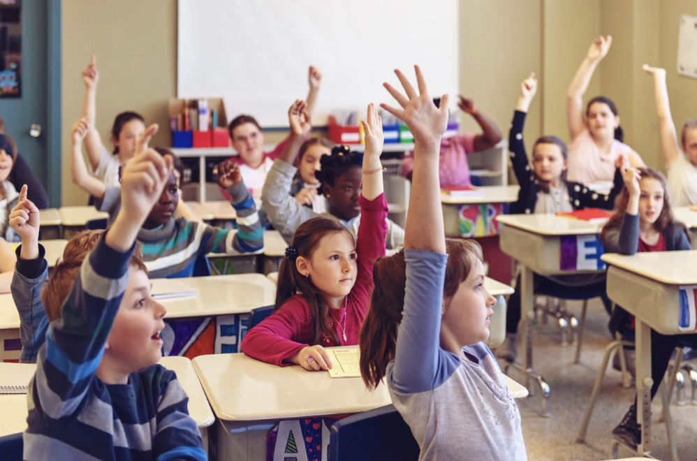 Photo of children with hands raised inside a classroom